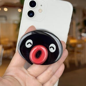 May include: A black and red phone grip with a cartoon face design. The grip is attached to a white smartphone. The cartoon face has a black background, white eyes, and a red mouth.