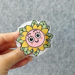 May include: A clear, circular sticker featuring a cartoon sunflower with a smiling face. The sunflower has yellow petals, green leaves, and a pink center. The sticker is held by a hand against a gray background.