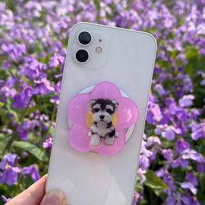 May include: A white smartphone with a pink flower-shaped phone grip featuring a puppy design. The puppy is black, white, and gray. The background is a field of purple flowers.