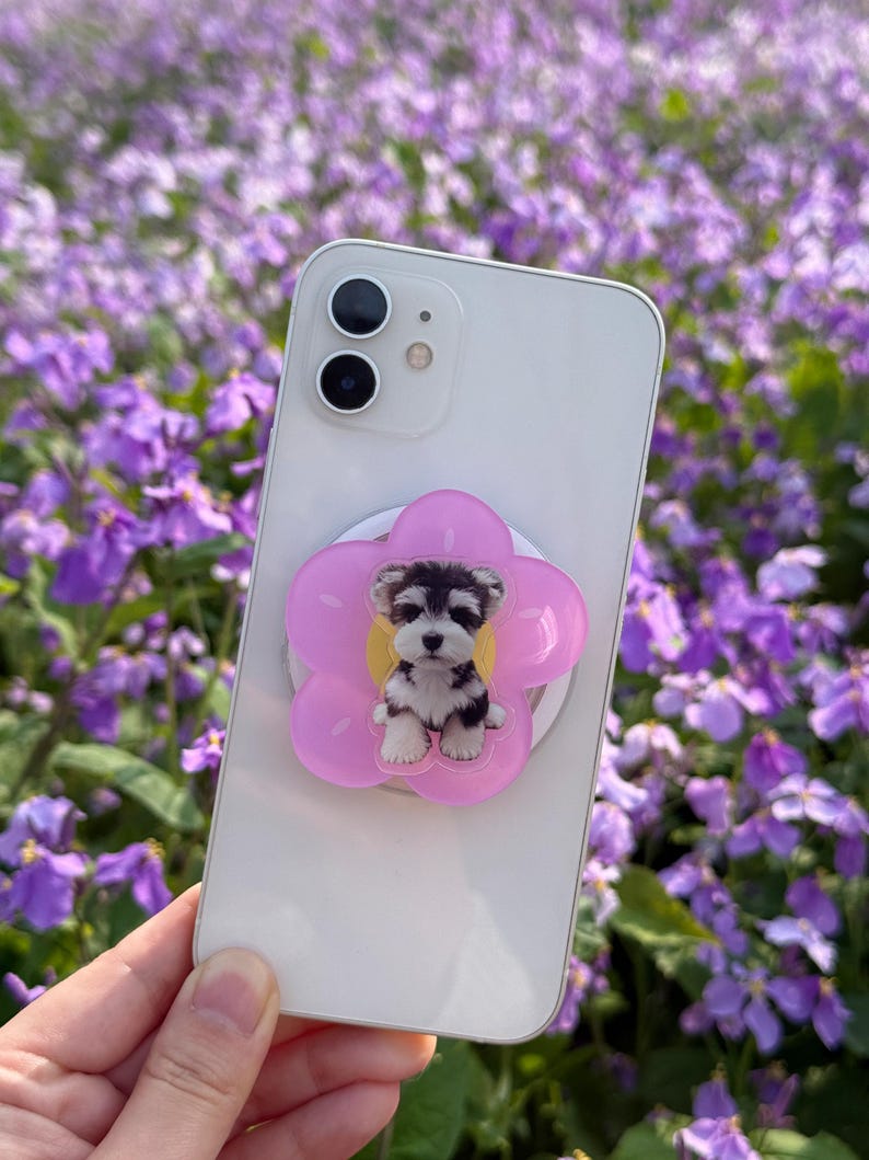 May include: A white smartphone with a pink flower-shaped phone grip featuring a puppy design. The puppy is black, white, and gray. The background is a field of purple flowers.