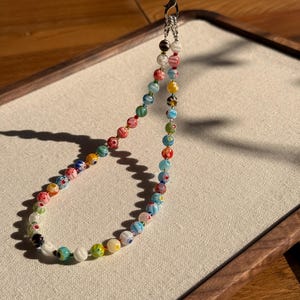 May include: A phone charm strap featuring a loop of multicoloured glass beads with intricate floral patterns. The beads are strung on a thin cord and attached to a silver-toned metal lobster clasp and a short black string loop for attachment.