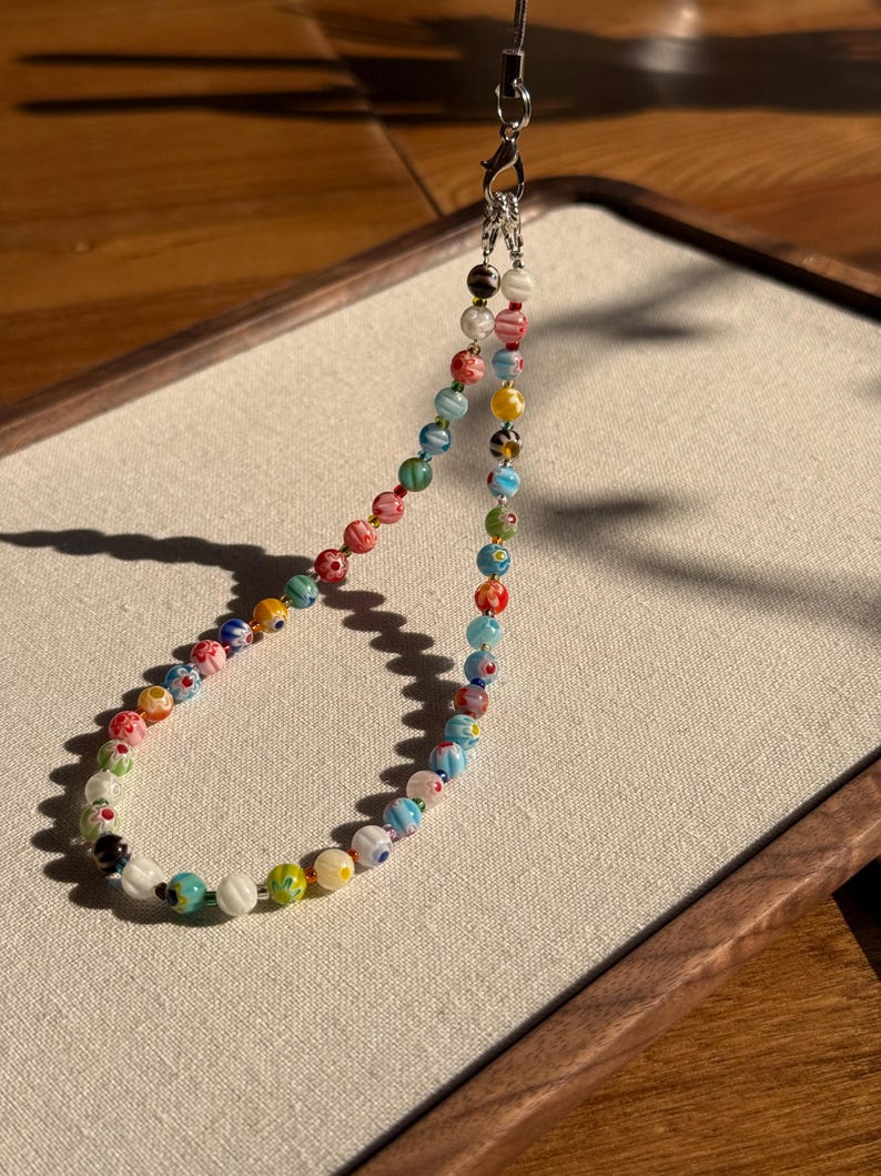 May include: A phone charm strap featuring a loop of multicoloured glass beads with intricate floral patterns. The beads are strung on a thin cord and attached to a silver-toned metal lobster clasp and a short black string loop for attachment.
