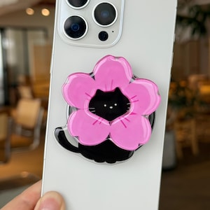 May include: A light gray smartphone with a pink flower-shaped phone grip featuring a black cat design. The phone grip is attached to the back of the phone. The phone has a triple-lens camera.