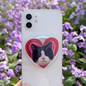 May include: A white smartphone with a pop-up grip featuring a black and white cat face inside a red heart design. The background is a field of purple flowers.