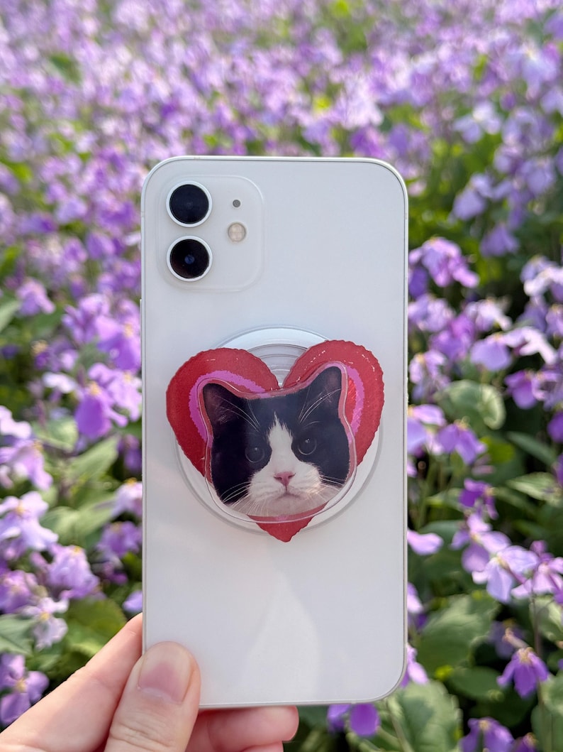 May include: A white smartphone with a pop-up grip featuring a black and white cat face inside a red heart design. The background is a field of purple flowers.