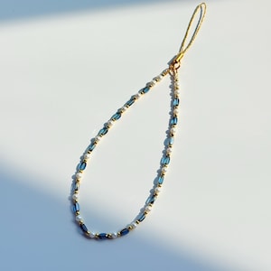 May include: A phone charm featuring a loop of alternating blue glass beads, small white pearls, and tiny gold-toned metallic spacers. The beaded strand is attached to a thin, shimmering gold-coloured cord loop for secure attachment to a mobile device.