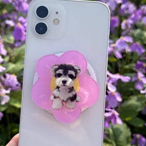 May include: A white smartphone with a pink flower-shaped phone grip featuring a black, white, and brown puppy. The phone is held in front of a background of purple flowers.