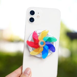May include: A white smartphone with a colorful pinwheel-shaped phone grip attached. The pinwheel features red, yellow, green, and blue blades. The phone has two camera lenses.