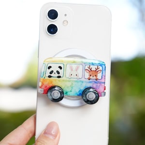 May include: A colorful phone grip featuring a rainbow-colored bus with a panda, rabbit, and deer inside. The bus has black wheels and is attached to a white phone. The background is blurred.