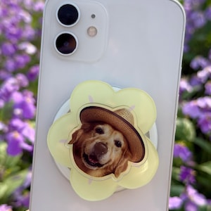 May include: A light-colored smartphone with a yellow flower-shaped phone grip featuring a smiling golden retriever wearing a straw hat. The phone grip is attached to the back of the phone.