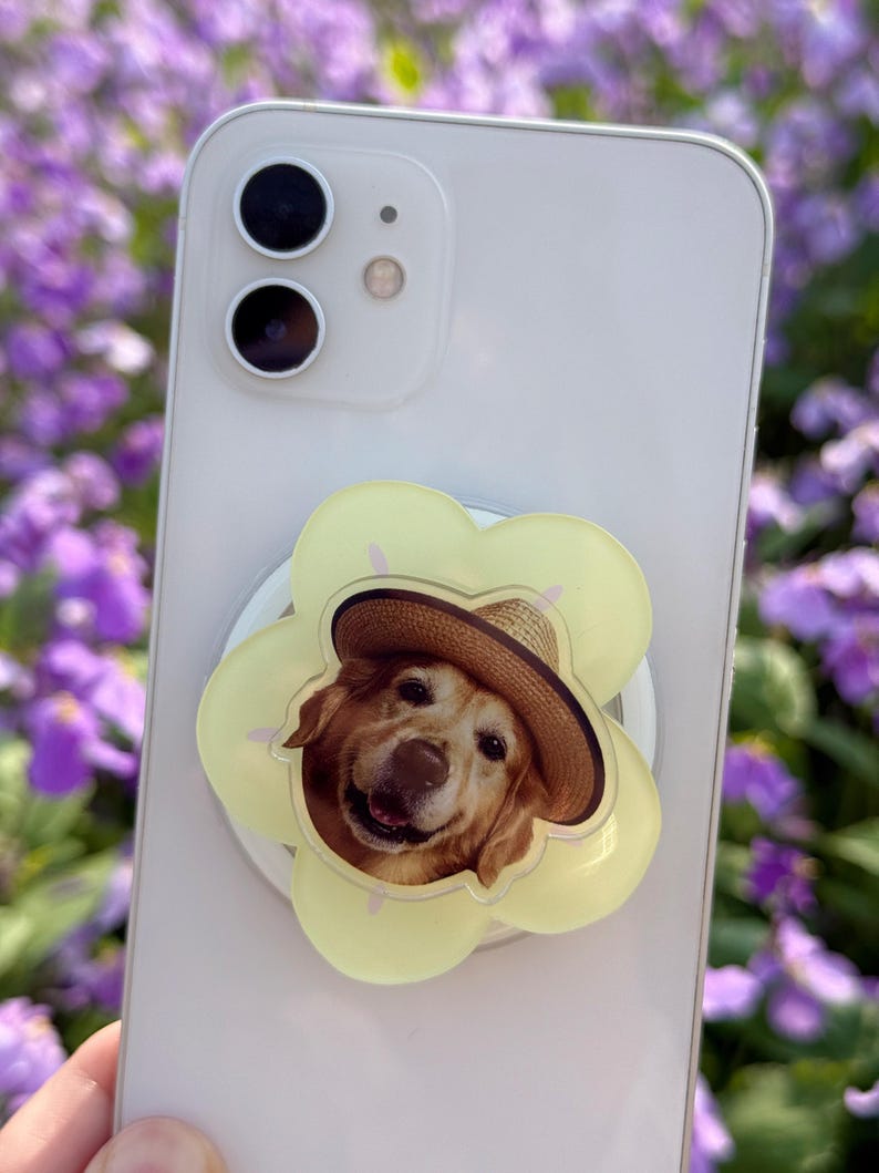 May include: A light-colored smartphone with a yellow flower-shaped phone grip featuring a smiling golden retriever wearing a straw hat. The phone grip is attached to the back of the phone.