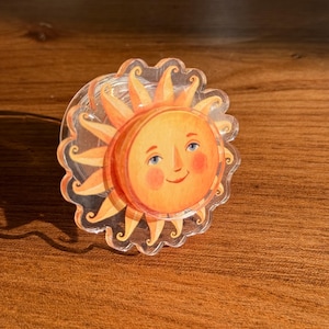May include: A clear, circular sun-shaped acrylic charm with a smiling face, rosy cheeks, and blue eyes. The sun has orange and yellow rays and a scalloped edge. The charm is on a wooden surface.