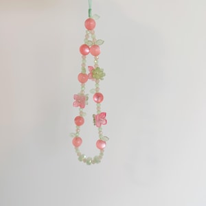 May include: A beaded phone charm featuring pink and green beads, with pink flower and fruit-shaped accents. The charm is suspended by a light green cord, creating a delicate and whimsical accessory.