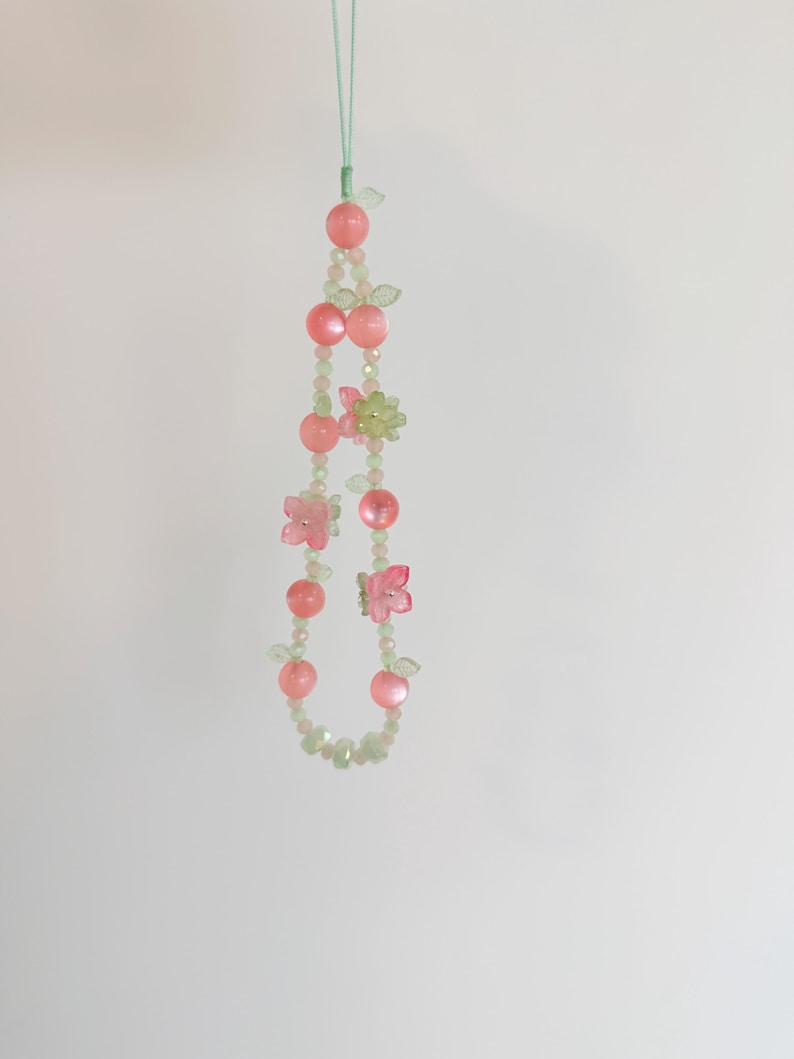 May include: A beaded phone charm featuring pink and green beads, with pink flower and fruit-shaped accents. The charm is suspended by a light green cord, creating a delicate and whimsical accessory.