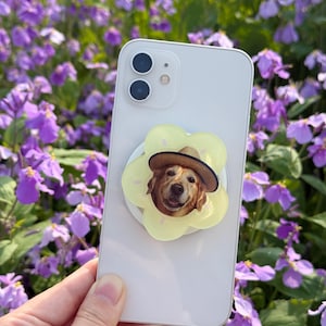 May include: A white smartphone with a yellow flower-shaped phone grip featuring a Golden Retriever wearing a straw hat. The phone is held in front of a background of purple flowers.