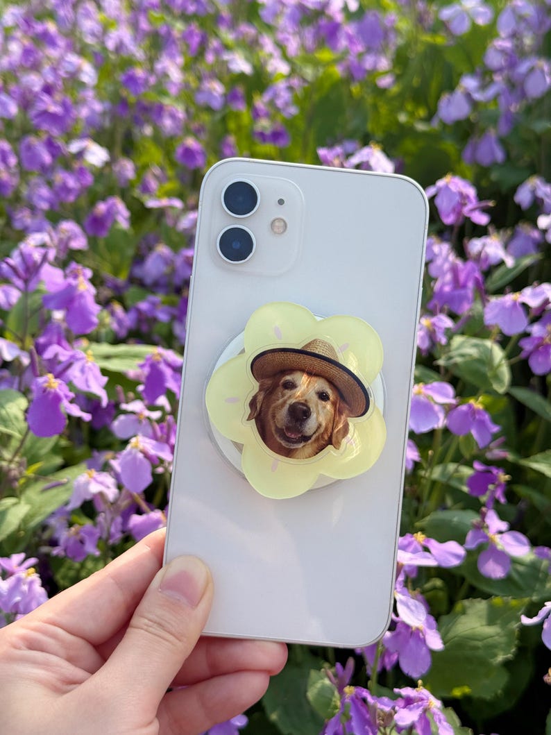 May include: A white smartphone with a yellow flower-shaped phone grip featuring a Golden Retriever wearing a straw hat. The phone is held in front of a background of purple flowers.