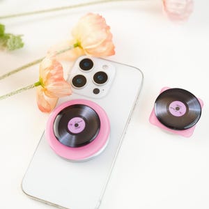 May include: A white smartphone with a pink pop socket featuring a vinyl record design. A second pop socket and a few peach-colored flowers are also visible. The pop sockets have a black record design with a pink center.