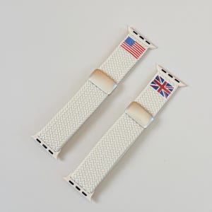 Op de afbeelding: Twee crèmekleurige gevlochten Apple Watch-bandjes. Eén bandje toont de Amerikaanse vlag en de andere de Union Jack. Beide bandjes hebben zilverkleurige sluitingen en connectoren.