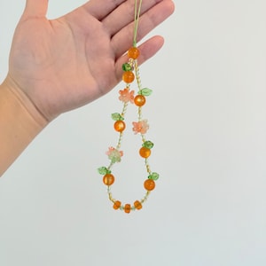 May include: A beaded necklace featuring orange and green beads, with small flower and leaf accents. The necklace is held up against a white background. The beads are arranged in a circular pattern, with a green cord.