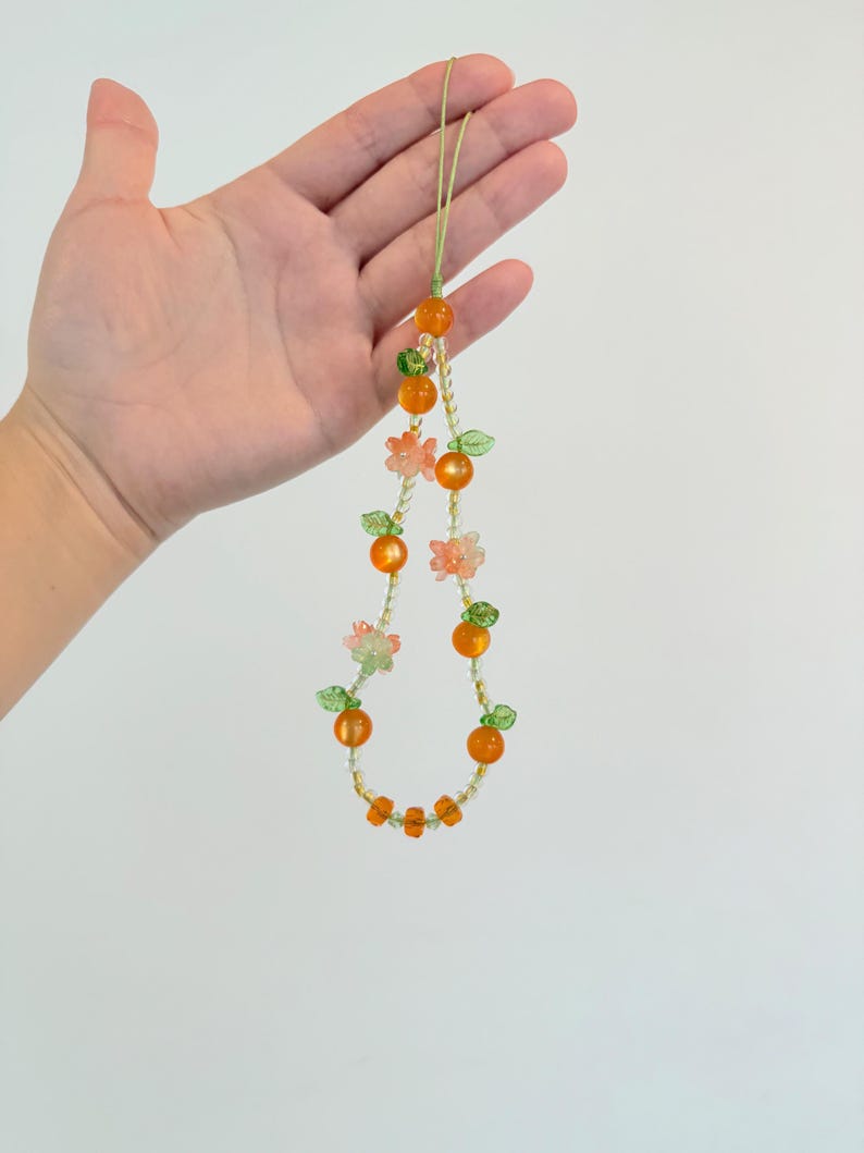 May include: A beaded necklace featuring orange and green beads, with small flower and leaf accents. The necklace is held up against a white background. The beads are arranged in a circular pattern, with a green cord.
