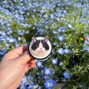 May include: A black and white cat face design on a round phone grip. The grip has a black and white striped border and is held against a background of blue flowers.