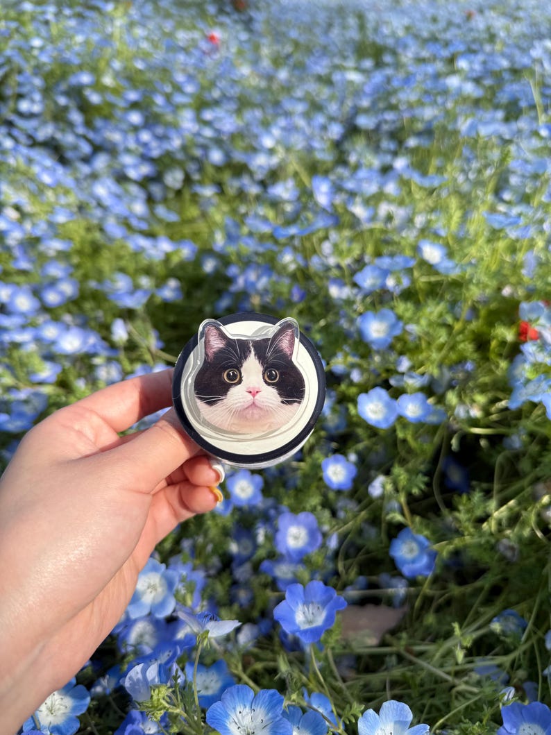 May include: A black and white cat face design on a round phone grip. The grip has a black and white striped border and is held against a background of blue flowers.
