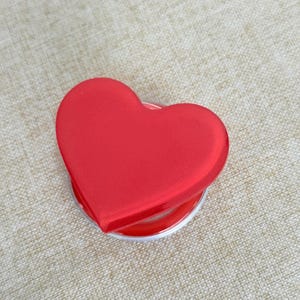 Peut inclure: Un support de téléphone en forme de cœur rouge avec une finition mate lisse. La surface supérieure est un cœur rouge vif et solide, tandis que la base est un composant circulaire en plastique translucide permettant à l'accessoire de se déployer.