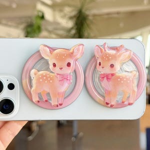 May include: Two pink and orange deer phone grips with a bow tie and white spots. The deer are set inside a pink circle. The phone grips are attached to a white phone.