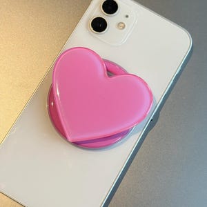 Op de afbeelding: Een witte smartphone met een roze hartvormige telefoonhouder bevestigd. De telefoonhouder heeft een levendige roze kleur en is gecentreerd op de achterkant van de telefoon. De telefoon heeft een zilveren frame en een camera met dubbele lens.
