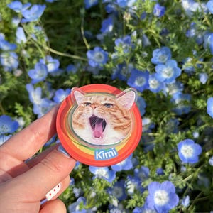 May include: A round, colorful phone grip with a cat's face in the center. The cat is orange and white and has its mouth open as if it is yawning. The phone grip has a rainbow design around the edge and the word "Kimi" written in white letters on the bottom.