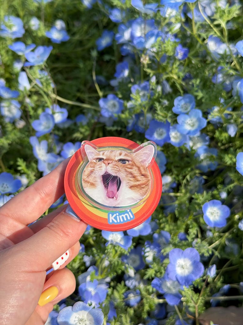 May include: A round, red cat-themed phone grip with a rainbow border. The image features an orange tabby cat with its mouth open. The name "Kimi" is printed in white on the bottom. The background is a field of blue flowers.