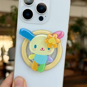 May include: A light blue smartphone with a colorful cartoon bunny phone grip. The bunny has blue, yellow, orange, green, and purple accents, and a yellow flower. The phone grip is attached to the back of the phone.