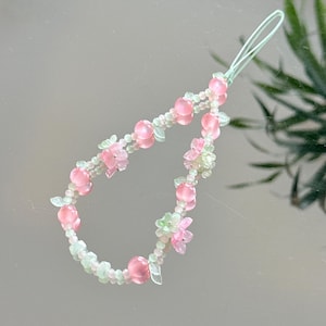 May include: A beaded phone charm featuring pink and clear beads, with small pink flower and leaf accents. The charm has a light green loop for attachment. The beads are arranged in a curved pattern.