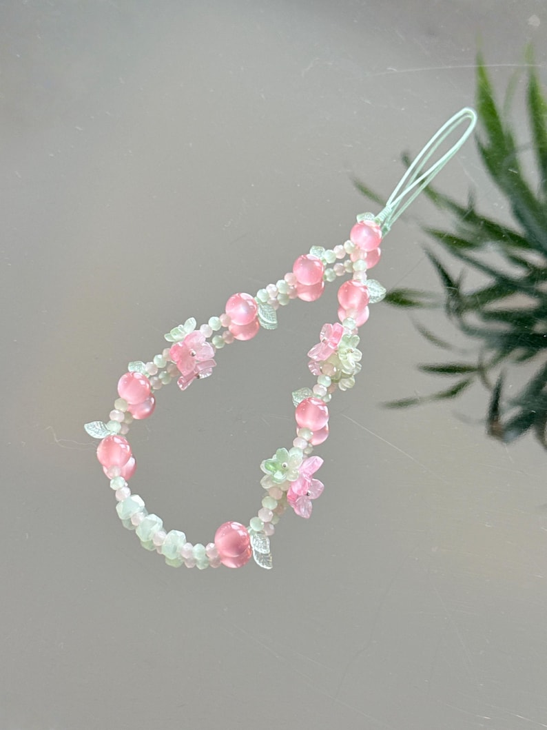 May include: A beaded phone charm featuring pink and clear beads, with small pink flower and leaf accents. The charm has a light green loop for attachment. The beads are arranged in a curved pattern.