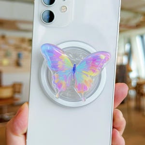May include: A white smartphone with a colorful butterfly phone grip attached. The butterfly has iridescent wings with shades of pink, yellow, and blue. The phone grip is round and clear.