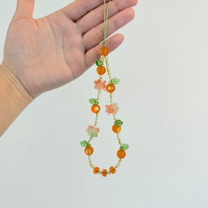May include: A beaded necklace featuring orange and clear beads, with small green leaf and pink flower accents. The necklace is held up by a light green string. The necklace has a fruit theme.