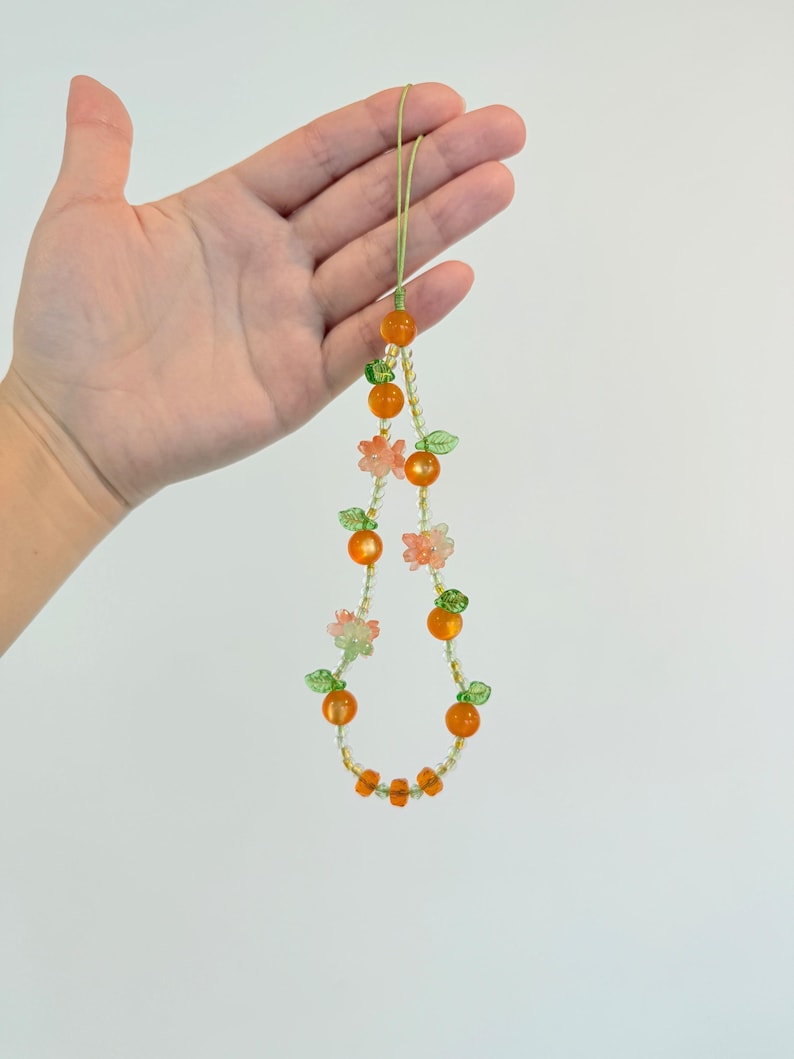 May include: A beaded necklace featuring orange and clear beads, with small green leaf and pink flower accents. The necklace is held up by a light green string. The necklace has a fruit theme.