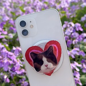 May include: A white smartphone with a heart-shaped phone grip featuring a cat's face. The grip has a red and pink striped border. The background is a field of purple flowers.