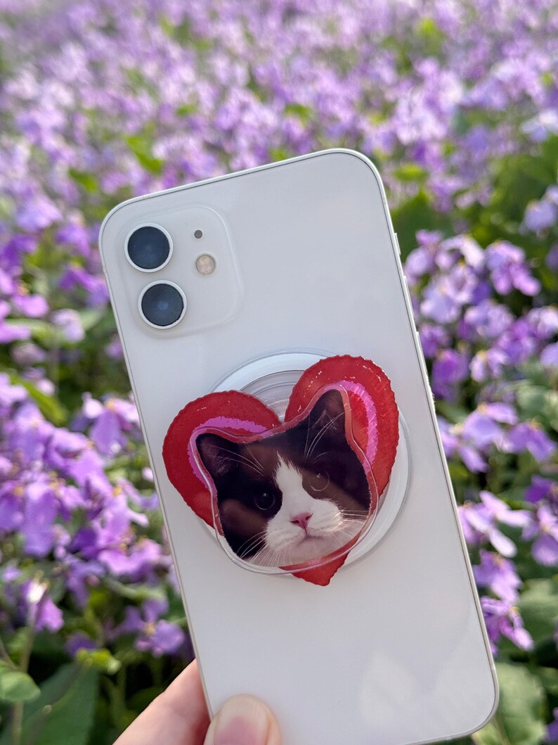May include: A white smartphone with a heart-shaped phone grip featuring a cat's face. The grip has a red and pink striped border. The background is a field of purple flowers.