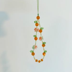 May include: A beaded phone charm featuring orange beads resembling oranges, interspersed with green leaf and pink flower beads. The charm is suspended from a green cord, creating a whimsical and colorful accessory.