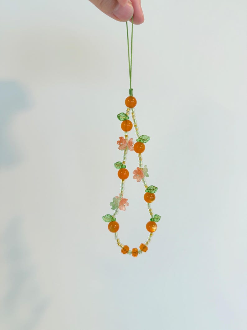 May include: A beaded phone charm featuring orange beads resembling oranges, interspersed with green leaf and pink flower beads. The charm is suspended from a green cord, creating a whimsical and colorful accessory.
