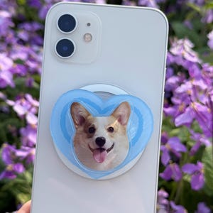 May include: A white smartphone with a light blue heart-shaped phone grip featuring a smiling corgi dog. The phone grip is attached to the back of the phone. The background is blurred with purple flowers.