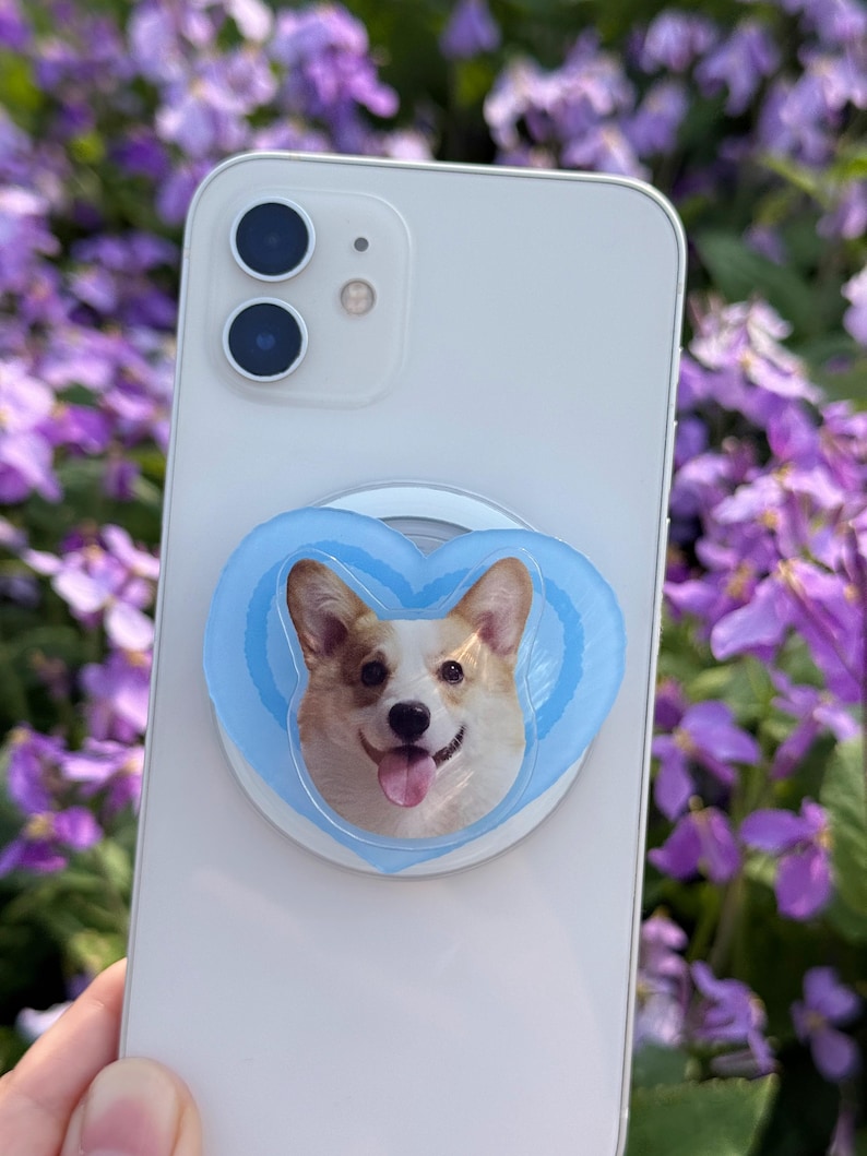 May include: A white smartphone with a light blue heart-shaped phone grip featuring a smiling corgi dog. The phone grip is attached to the back of the phone. The background is blurred with purple flowers.