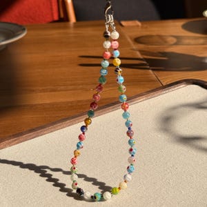 May include: A beaded lanyard featuring a variety of small, round glass beads in multiple colours including red, blue, yellow, green, and white. The beads display diverse patterns such as stripes, swirls, and floral motifs, connected by a silver-toned metal clasp.