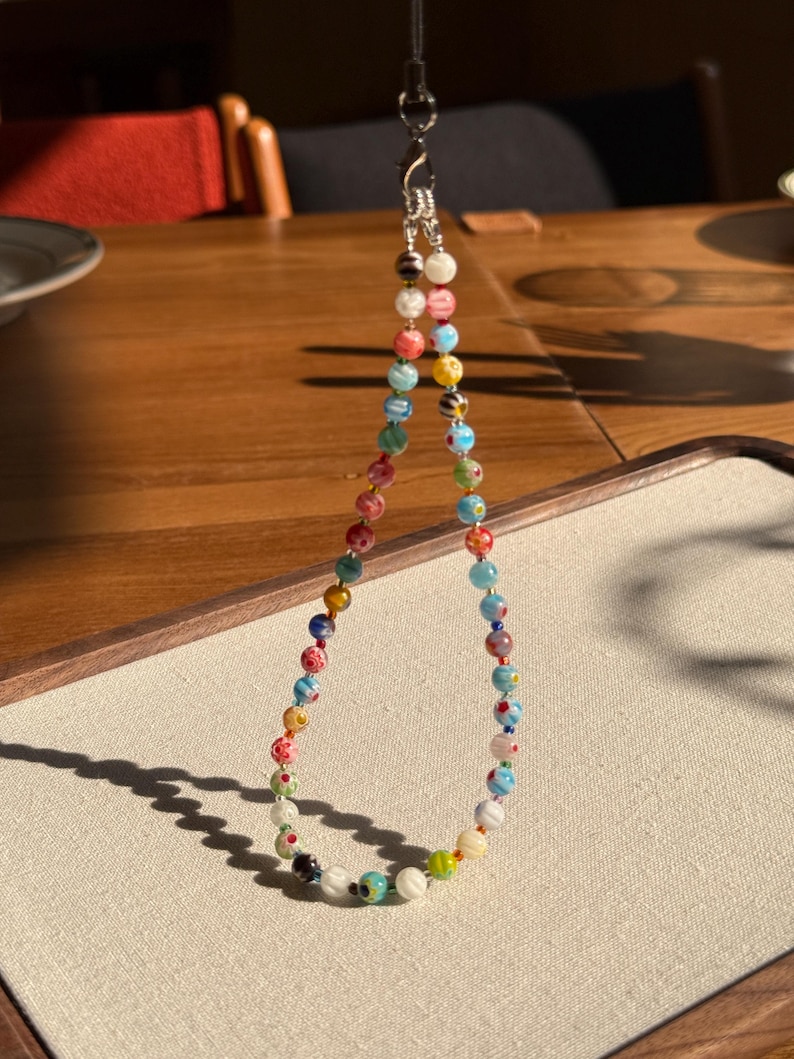 May include: A beaded lanyard featuring a variety of small, round glass beads in multiple colours including red, blue, yellow, green, and white. The beads display diverse patterns such as stripes, swirls, and floral motifs, connected by a silver-toned metal clasp.