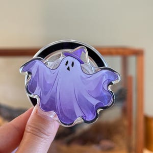 Op de afbeelding: Een paars spook met een heksenhoed, een Halloween-telefoonhouder. Het spook heeft een lachend gezicht en staat tegen een transparante achtergrond met een zwarte ring. De houder wordt door een hand vastgehouden.