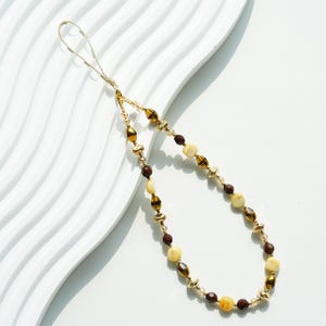 May include: A beaded phone charm featuring a loop of gold-toned cord, interspersed with faceted dark brown beads, smooth cream-colored beads, and small metallic gold spacers arranged in a repeating pattern to form a decorative loop accessory.