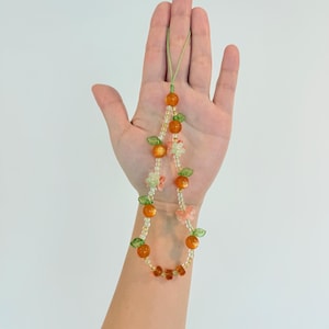 May include: A beaded phone charm featuring orange-colored beads, small green leaf accents, and clear beads. The charm also includes small flower and butterfly accents. The charm is held up against a white background.