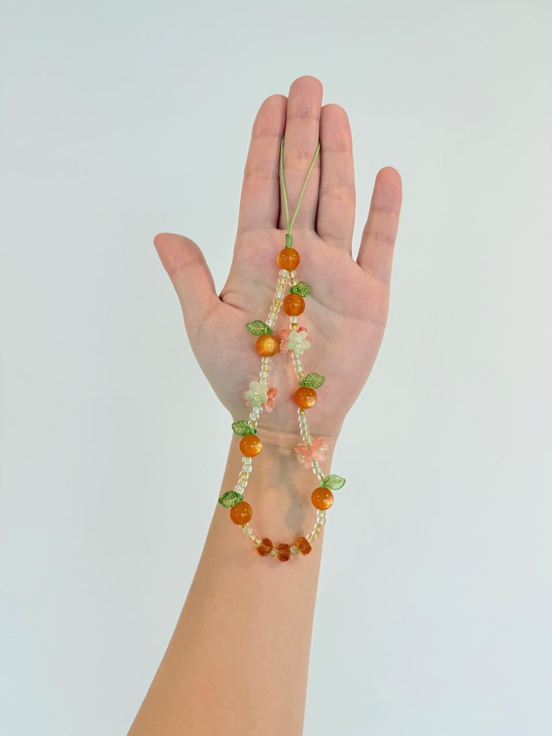 May include: A beaded phone charm featuring orange-colored beads, small green leaf accents, and clear beads. The charm also includes small flower and butterfly accents. The charm is held up against a white background.