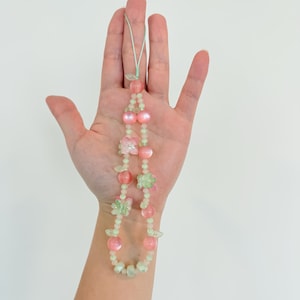 May include: A beaded necklace featuring pink and green beads, with flower and leaf accents. The necklace is held against a white background. The beads are a mix of round and flower-shaped, with a light green cord.